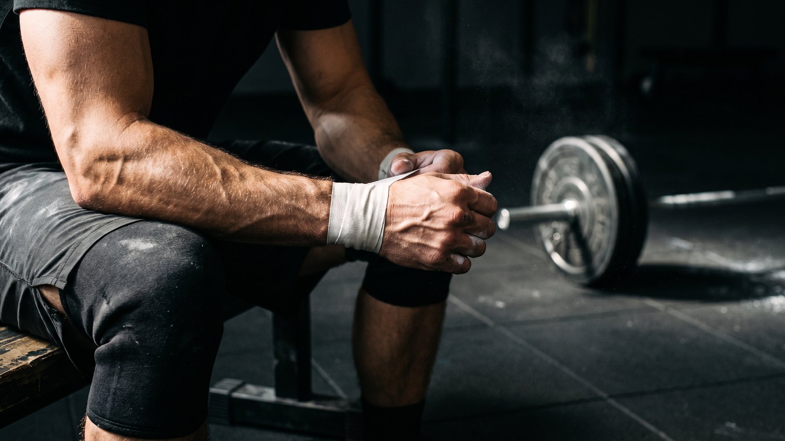 A man's hands taping a wrist on a gym bench, with a loaded barbell and chalk dust behind him under hard side light.